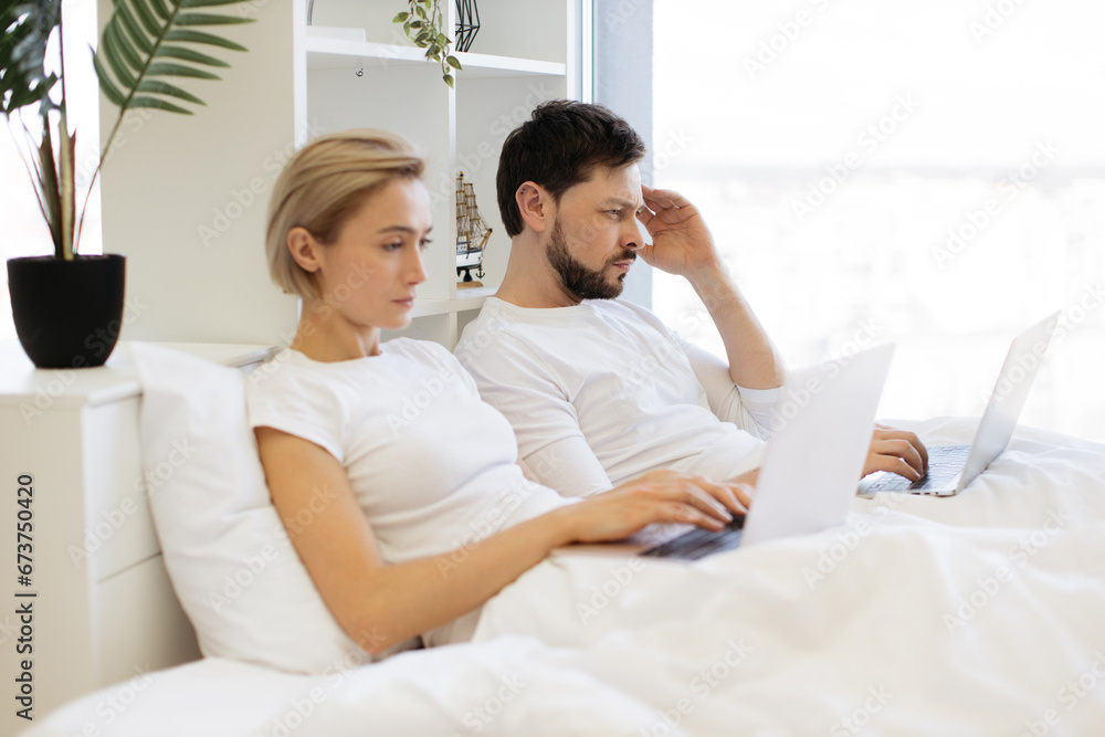 Attractive bearded caucasian man and pretty woman using digital devices while leaning against soft pillows in bed of studio room. Busy spouses doing their own business without disturbing each other. Attractive bearded caucasian man and pretty woman using digital devices while leaning against soft pillows in bed of studio room. Busy spouses doing their own business without disturbing each other.
