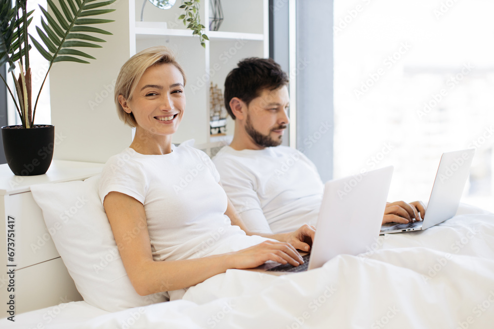 Handsome bearded man making notes on laptop while smiling woman activating computer on her knees. Caucasian married couple doing efficient distant work while staying in bed at home. Handsome bearded man making notes on laptop while smiling woman activating computer on her knees. Caucasian married couple doing efficient distant work while staying in bed at home.
