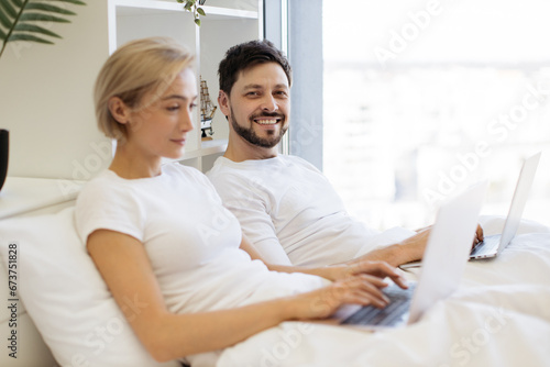 Smiling caucasian male scrolling through webpages on laptop in bed while busy lady typing on portable computer beside him. Mature people in relationships spending quality time together at home.