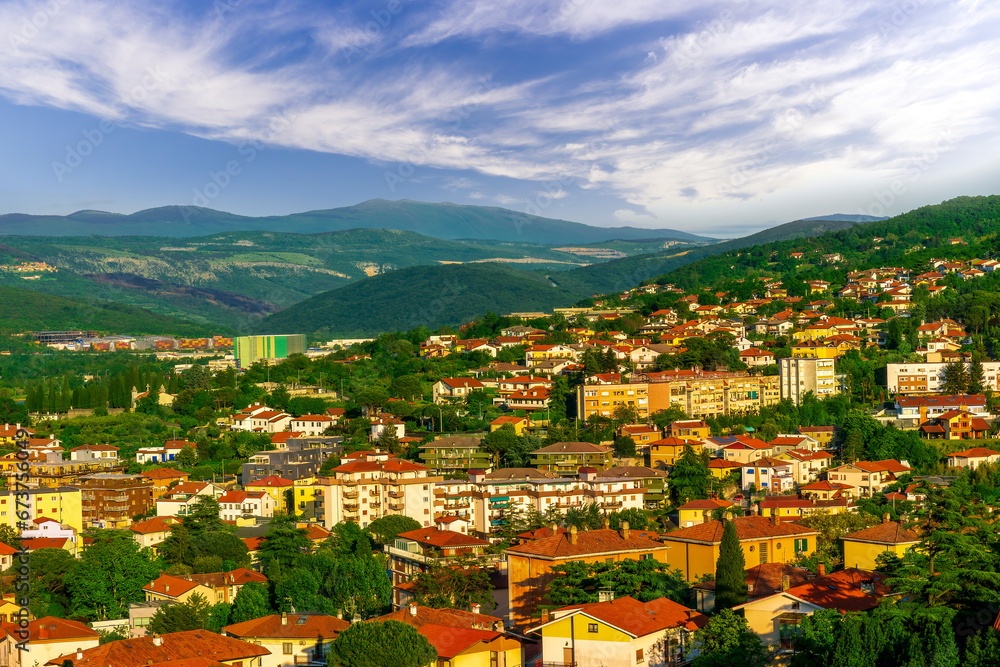 panoramic landscape view of green summer city with yellow houses, green forest and mountains above and amazing hills and cloudy sky on background