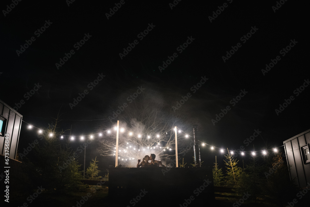 Couple enjoying hot tub at night outdoors. Winter holidays in mountains, hot water treatments concept. Man and woman swims in hot bath while resting open air. Glowing garlands and light bulbs. - obrazy, fototapety, plakaty 
