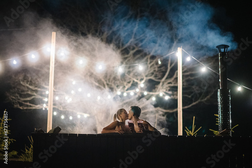 Couple enjoying hot tub at night outdoors. Winter holidays in mountains. Family relax in thermal spa open air. Man and woman swims in hot bath while resting. Glowing garlands and light bulbs. photo