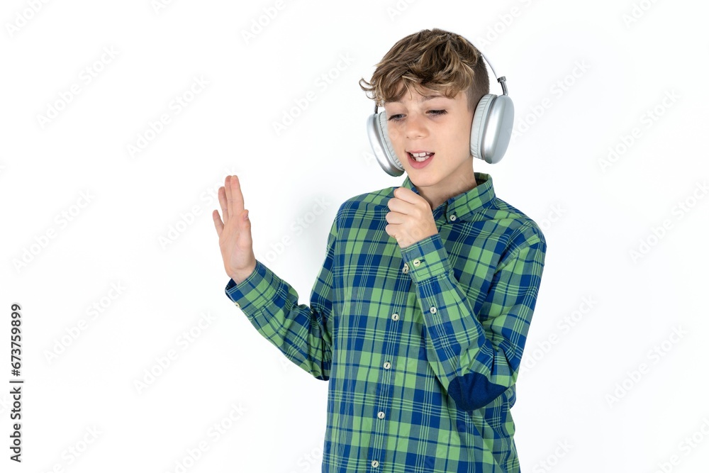 Happy Caucasian teen boy wearing plaid shirt sings favourite song keeps hand near mouth as if microphone wears wireless headphones, listens music Happy Caucasian teen boy wearing plaid shirt sings favourite song keeps hand near mouth as if microphone wears wireless headphones, listens music