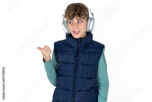  Caucasian teen boy wearing vest jacket  listens audio track via wireless headphones points thumb away advertises copy space photo