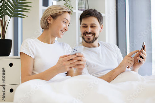 Happy caucasian couple holding cellphones in hands while lounging in soft bed all morning indoors. Attractive man and woman browsing internet using mobile application in digital devices.
