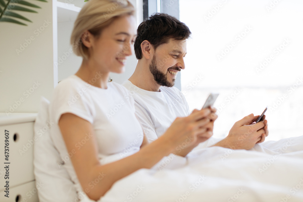 Serene caucasian family of two using mobile phones while lazing in soft bed during daytime at home. Confident adult man and woman reading online article paying attention to details. Serene caucasian family of two using mobile phones while lazing in soft bed during daytime at home. Confident adult man and woman reading online article paying attention to details.