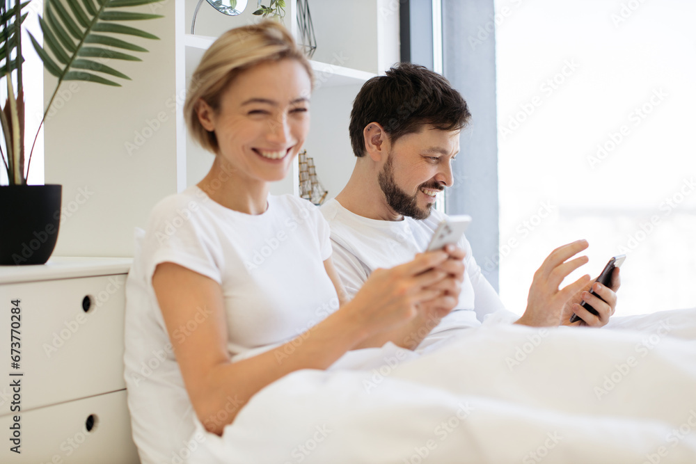 Serene caucasian family of two using mobile phones while lazing in soft bed during daytime at home. Confident adult man and woman reading online article paying attention to details. Serene caucasian family of two using mobile phones while lazing in soft bed during daytime at home. Confident adult man and woman reading online article paying attention to details.