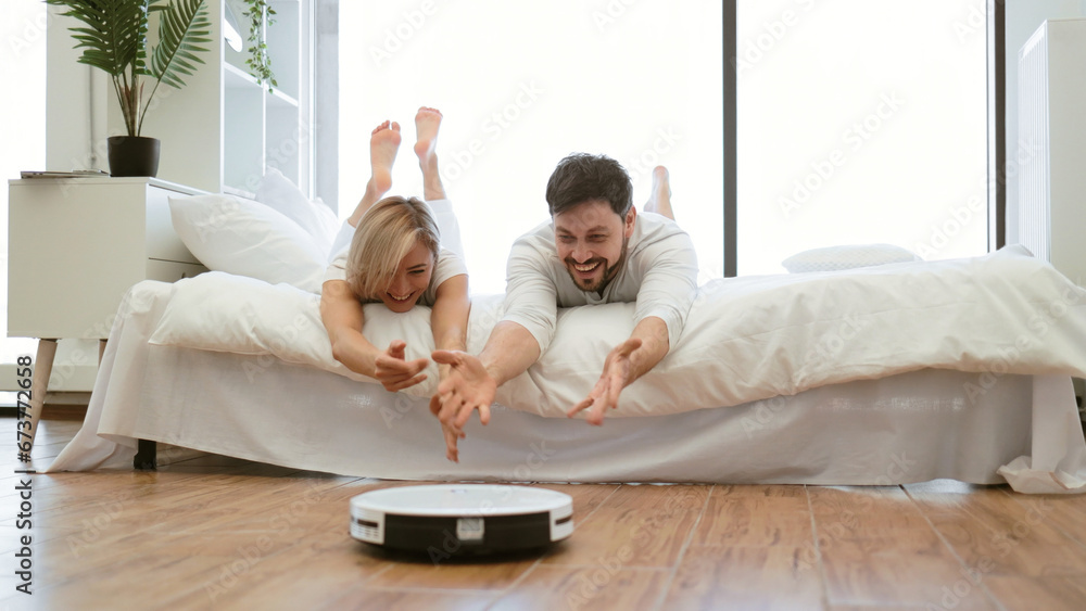 Smiling wife and husband having fun playing with smart cleaning robot while resting on bed at home. Family man and woman creating love bond by spending time together while spending time together. Smiling wife and husband having fun playing with smart cleaning robot while resting on bed at home. Family man and woman creating love bond by spending time together while spending time together.