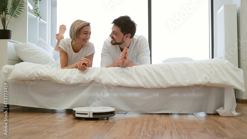 Robot vacuum carrying out scheduled cleaning task while male and female owners lying resting on bed. Smart device saving energy from tedious home cleaning work by using advanced technologies.