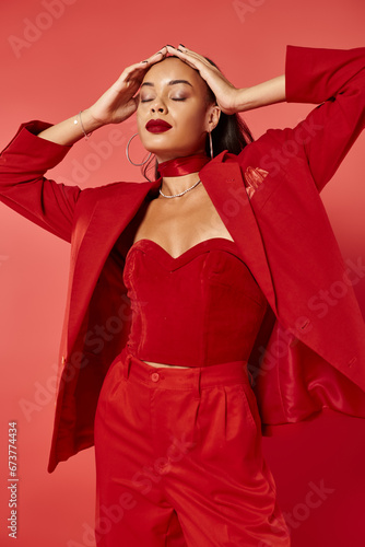 attractive african american woman with ponytail posing in suit with hand on head on red backdrop