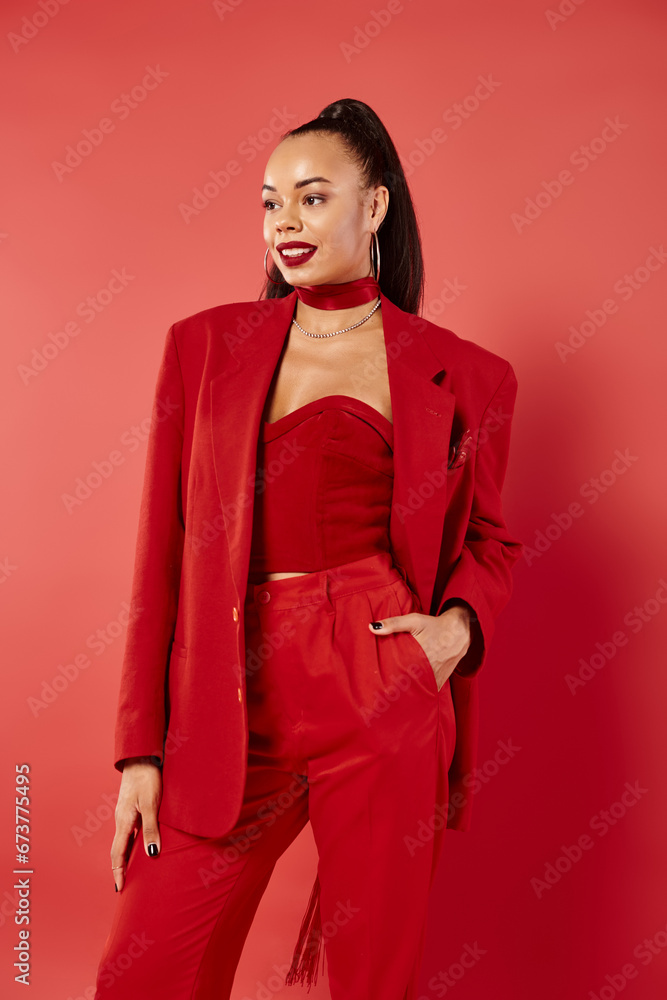 happy african american woman with ponytail posing with hand in pocket of pants suit on red backdrop happy african american woman with ponytail posing with hand in pocket of pants suit on red backdrop