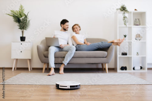 Automatic robot vacuum cleaning wooden floor while happy young couple resting on background. Two caucasians in love spending time for watching movie instead of households.
