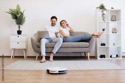 Young caucasian couple in casual clothes relaxing on comfy couch with portable laptop in hands. Family of two watching movie while wireless robot vacuuming wooden floor.