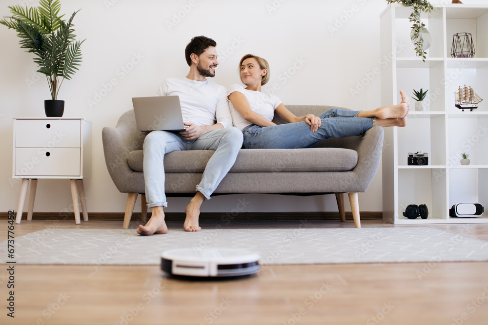 Young caucasian couple in casual clothes relaxing on comfy couch with portable laptop in hands. Family of two watching movie while wireless robot vacuuming wooden floor.