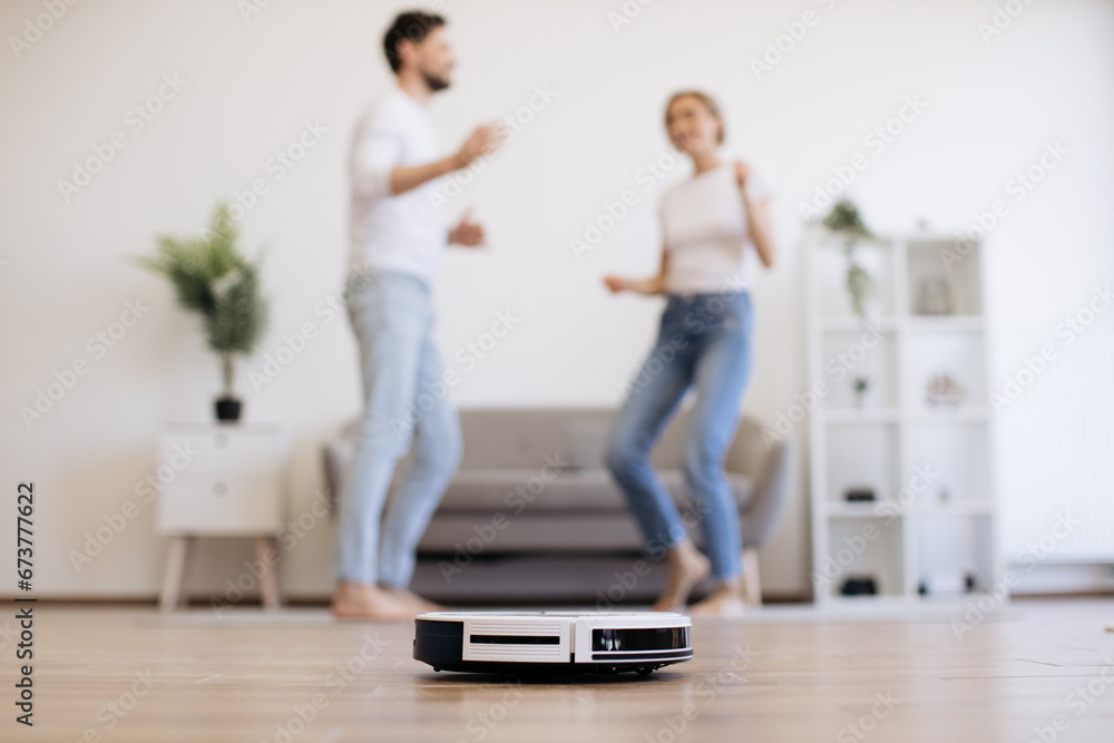 Cheerful lady in casual clothes dancing with husband during home dancing party with working robot cleaner on floor. Happy caucasian family man and his wife entertaining while doing home care.