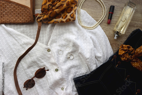 White blouse, denim skirt, brown shoes, vintage bag, leopard print scarf, pearl necklace, scrunchie, silver rings, perfume and lipstick on wood background. Retro, elegant outfit. Top view.