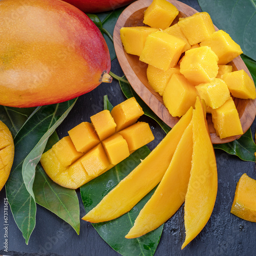 Ripe mango fruits with slices and mango leaves on a gray stone table.