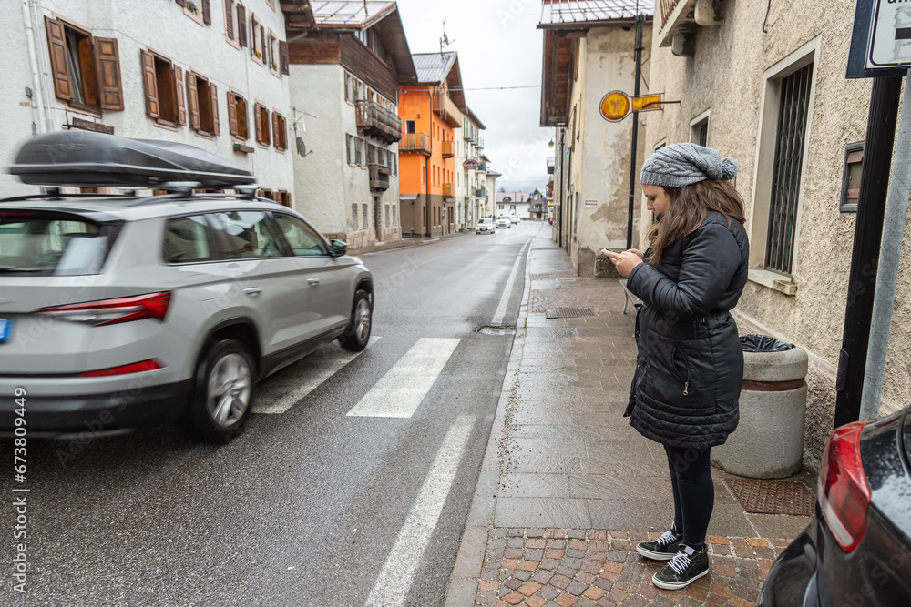 Female tourist in winter black clothes standing on sidewalk looking at his phone while cars ride on road; Venas di Cadore village; Dolomites; Italy