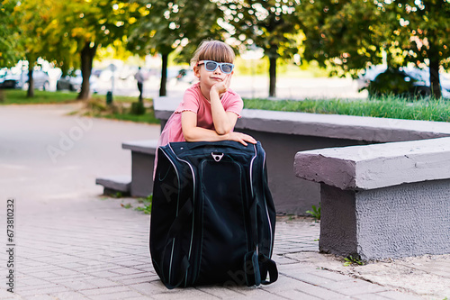 portrait of a little charming five year old girl resting next to a large black suitcase on a sunny day. A little girl stands with her luggage, waiting for transport. Travel, vacation, adventure