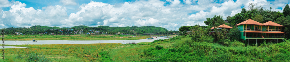 Panoramic view of a lush riverside village with traditional houses amid verdant hills under a dynamic sky. Panoramic view of a lush riverside village with traditional houses amid verdant hills under a dynamic sky.