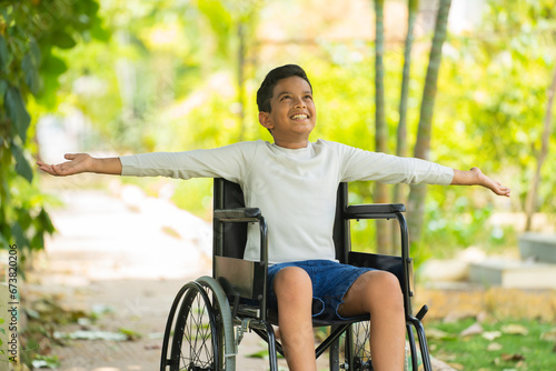 Happy indian kid with disability feeling fresh air by stretching hands sitting on wheelchair at park - concept of rehabilitation, health recovery and freedom.
