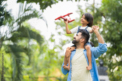 Happy smiling indian girl kid playing with airplane toy by sitting on father shoulder at park - concept of freedom, family support and togetherness photo