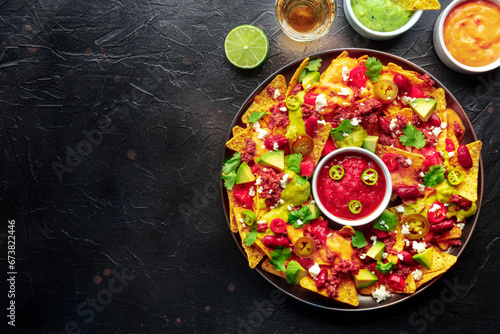 Loaded nachos. Mexican nacho chips with beef, overhead flat lay shot with guacamole sauce, cheese salsa, tequila drinks, limes, on a black slate background with copy space