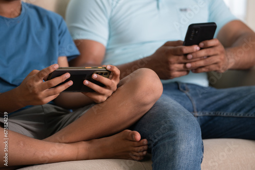 Close up shot of indian father and son hands busing using on there mobile phones while sitting on sofa at home - concept of technology addiction, cyberspace and disconnected