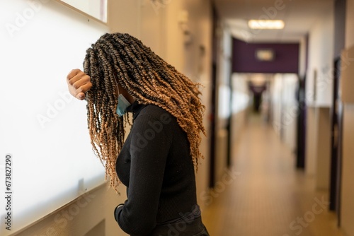 Young female with dreadlocks wearing a face covering leaning on a window in a corridor photo