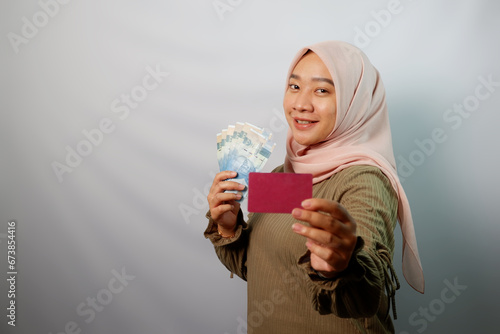Happy beautiful Asian Muslim woman holding paper money and  showing credit card isolated on white background. ​ photo
