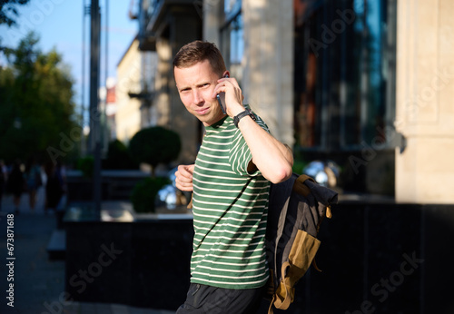 Young sporty man in a striped T-shirt walking on the street with a smartphone in hand and a trendy hipster's backpack slung over his shoulder, enjoying a leisurely urban outing.