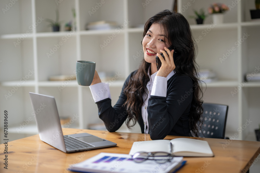 Freelance Asian woman talking on the phone and sipping coffee while working at the desk. Freelance Asian woman talking on the phone and sipping coffee while working at the desk.