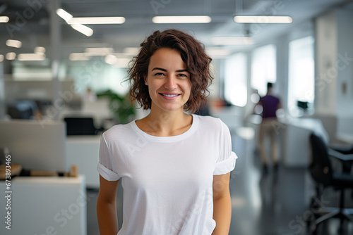 A young white brunette woman with a friendly smile, white teeth, curly hair, and a blank white t-shirt standing against a blurred office background. Mock-up for design. Blank template. photo