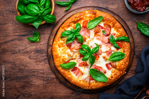 Homemade pizza with pancetta, mozzarella cheese, spicy sauce and green basil leaves on old wooden table background, top view