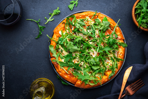Hot homemade pizza with blue cheese, ham, pesto sauce and fresh arugula, black table background, top view