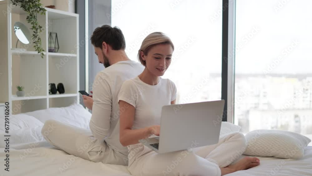 Smiling woman reading social updates using smartphone and serene man shopping online. Side view of caucasian couple in love working on modern gadgets while sitting back to back on bed. Smiling woman reading social updates using smartphone and serene man shopping online. Side view of caucasian couple in love working on modern gadgets while sitting back to back on bed.