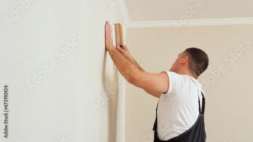 Vinyl wallpapers squeegee tool in use by professional interior renovation worker. Young man with dark hair in a uniform glueing beige wallpaper at home.