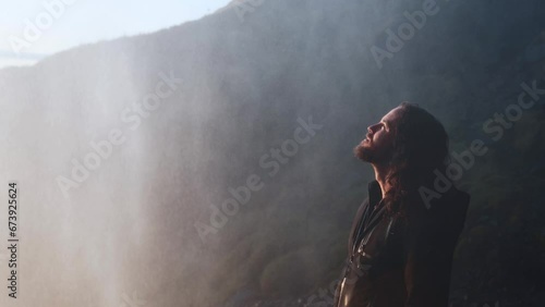 Man near big waterfall. Explorer with beard and long hair admires nature. Dramatic sunset light, hiker in cool waterfall mist. Freedom, northern lands, cold wilderness.