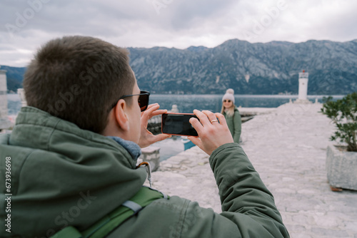 Husband takes a picture of his wife on a pier with a lighthouse in the background. Back view