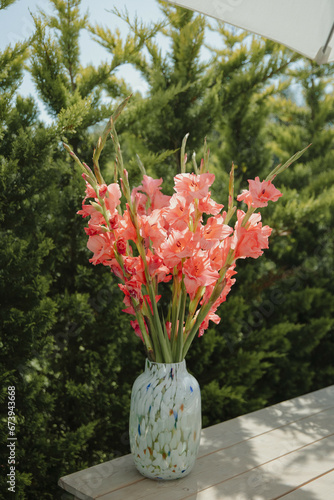 Vibrant pink gladiolus flowers in a decorative vase on a wooden table, set against lush green foliage.
