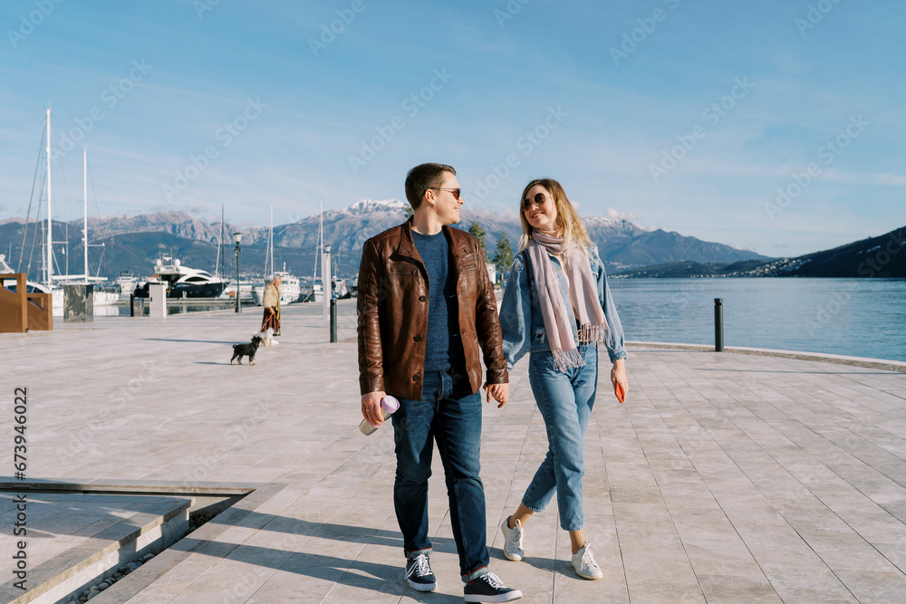 Man and woman walk holding hands and looking at each other along the pier by the sea