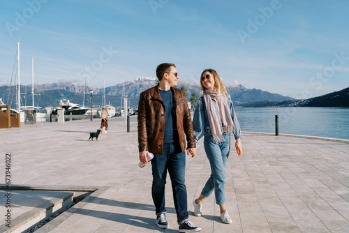 Man and woman walk holding hands and looking at each other along the pier by the sea