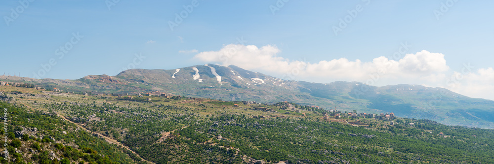 A village sits on a hill ridge across a rocky landscape, with Mount Lebanon mountain range, snow-covered in spring, a popular hiking destination in Lebanon, Middle East