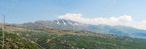 A village sits on a hill ridge across a rocky landscape, with Mount Lebanon mountain range, snow-covered in spring, a popular hiking destination in Lebanon, Middle East photo