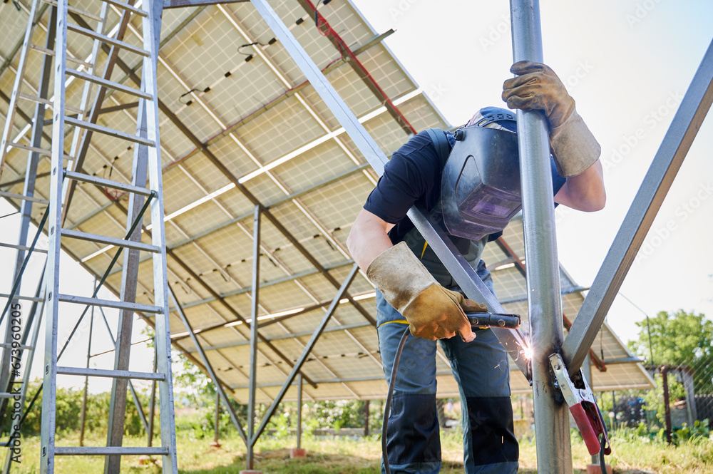 Welder welding metal beam with welding machine for installing solar panels. Worker in protective welding helmet wearing workwear, working on solar station on blurred background. Welder welding metal beam with welding machine for installing solar panels. Worker in protective welding helmet wearing workwear, working on solar station on blurred background.