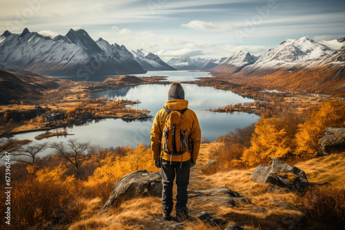 A lone hiker gazes over a serene lake reflecting autumnal golden hues and snow-dusted mountain peaks, capturing a moment of tranquil beauty. © apratim