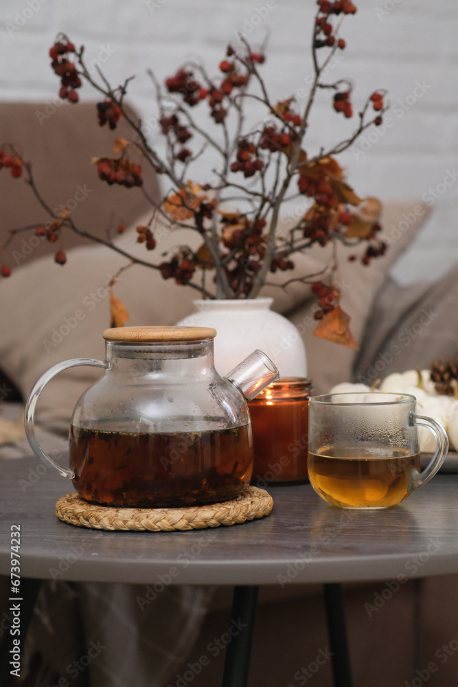 Autumn home design - hot teapot with tea, glass of tea, burning candle, vase with dry branches on sofa background in cozy living room.