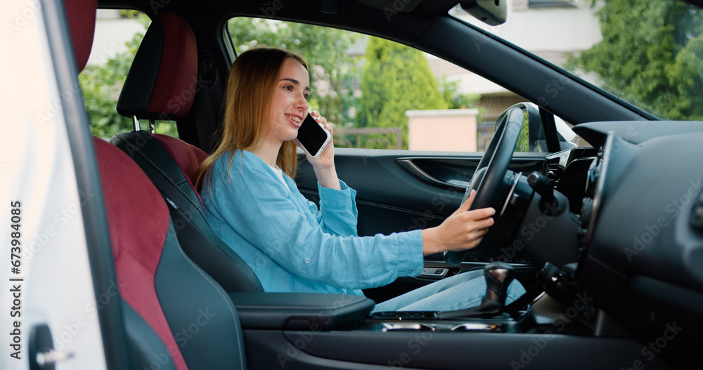 Naklejka premium Attractive ginger woman talks on smartphone while sits sits in car. Girl driver calling smartphone in modern automobile standing at sunny shore. Attractive blonde speaking on telephone inside auto.