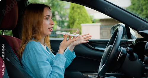 Fotografie Focused blonde lady sitting behind the wheel of car when in recording voice message on smartphone in business car