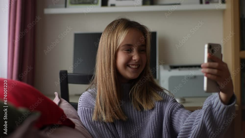 Portrait of cheerful adorable child girl holding and using mobile phone, having video call, showing thumb up, like gesture, sitting on couch in living room with modern interior, slow motion, closeup. Portrait of cheerful adorable child girl holding and using mobile phone, having video call, showing thumb up, like gesture, sitting on couch in living room with modern interior, slow motion, closeup.